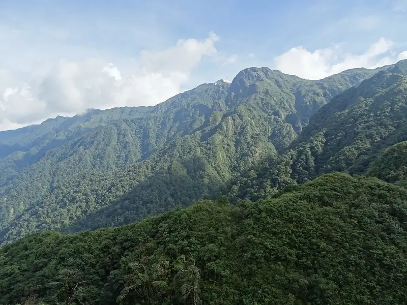Fansipan's green ridges under a blue sky with white cloud, seen across the forest from the trail